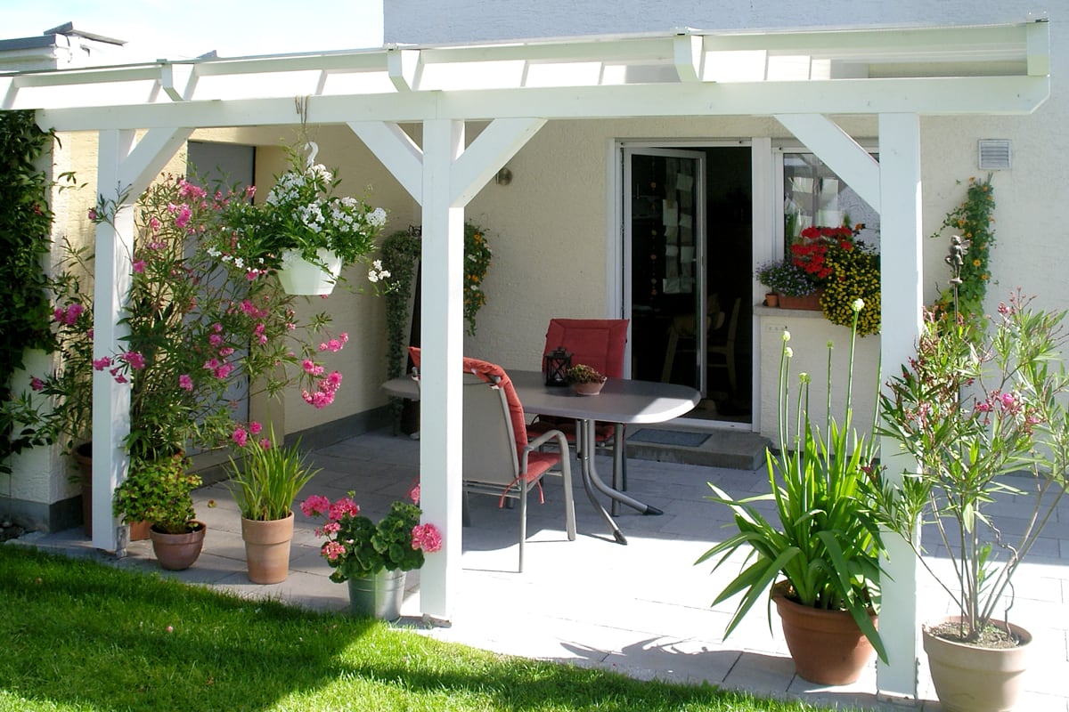 Eine weiß gestrichene Terrassenüberdachung aus Holz schützt eine kleine Sitzgruppe und ist von zahlreichen Blumentöpfen und Hängepflanzen umgeben.